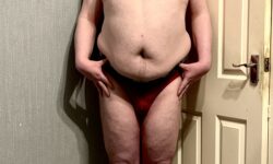 A tall bearded man with his hands on his hips wearing a pair of shiny burgundy red briefs with a dark burgundy red waistband that has Box Menswear written in bold black writing on it stood against a white wooden door.