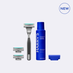 A tall cylindrical gradient black and dark silver metal razor handle that has a bright silver razor blade attached to the top of it next to two rectangular silver metal razor blades, a tall cylindrical dark blue metallic spray can that has Harry’s Sensitive Skin Foaming Shave Gel written in bright white writing on it, and a dark blue rubber shaving blade protector, on a light grey background.
