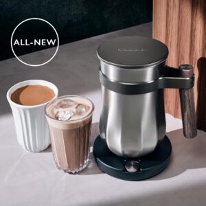 A large rectangular stainless steal pewter metal hot chocolate machine with a dark wooden handle on top of a shiny black base that has Hotel Chocolat engraved on it next to a tall cylindrical glass full of some creamy brown liquid and several clear ice cubes, and a short cylindrical bright white ceramic cup full of some dark brown liquid on a light wooden worktop, on a white background.
