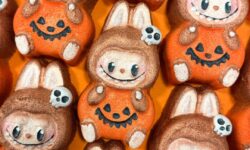 A pile of some medium bright orange Labubu Doll shaped Bath Bombs wearing a pumpkin costume and holding a small bright white skull shaped accessory that all have some big wide black eyes, some rosy pink cheeks, and some wide bright white smiles engraved on them, on a sparkly orange background.