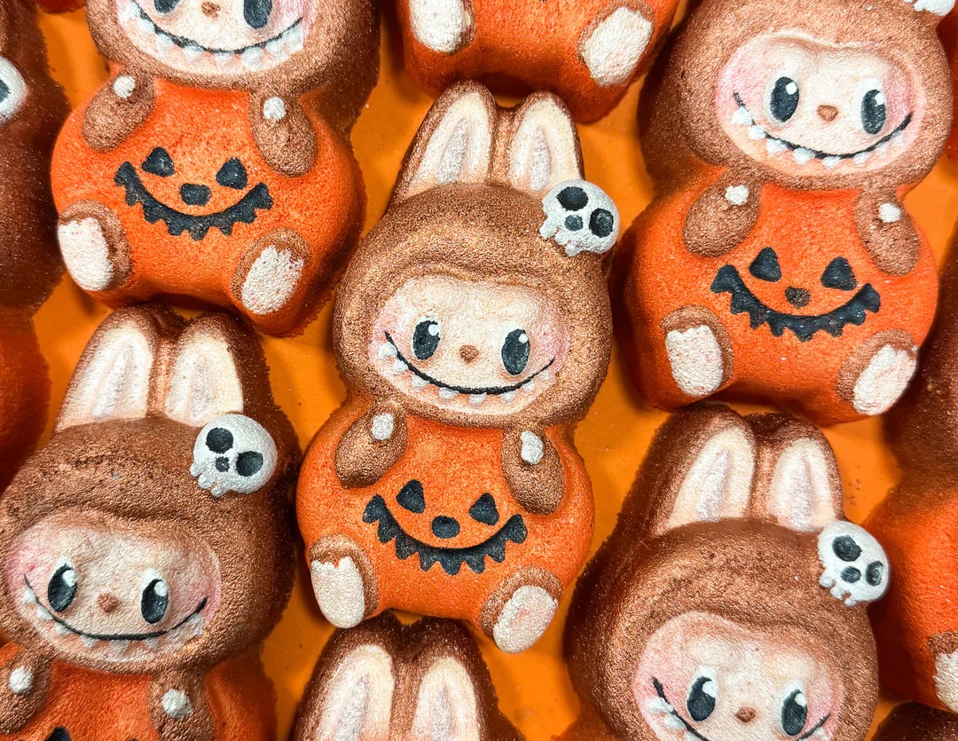 I Am The Owner Of The One And Only 24-Carat Gold Labumpkin in the World | Tales Of Toria Halloween 2025 Range A pile of some medium bright orange Labubu Doll shaped Bath Bombs wearing a pumpkin costume and holding a small bright white skull shaped accessory that all have some big wide black eyes, some rosy pink cheeks, and some wide bright white smiles engraved on them, on a sparkly orange background.