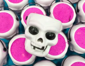 A pile of some bright white plastic skull shaped Bath Bomb Buckets full of some bright pink bath bomb powder that all have some black eye sockets and a large toothy grin engraved on them, on a dark blue background.