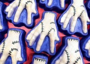 A pile of some large creamy beige monster’s severed hand shaped bath bombs that all have some dark blue wrists, some black stitches all over them, and some dark blue outlines around them, on a dark red background.