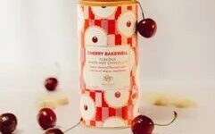 A tall cylindrical gradient bright pink, red and white geometric patterned cardboard tube with a dark blue plastic lid and a dark blue label that has Whittard of Chelsea Cherry Bakewell Flavour White Hot Chocolate written in bright gold writing on it next to some small circular golden brown bakewell tarts, some pieces of white chocolate and some small bright red cherries, on a white background.
