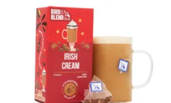 A medium bright red square cardboard box that has Bird & Blend Irish Cream Tea Bag Gift written in bright white writing and some pictures of some creamy white topped drinks and some pieces of dark brown chocolate on it next to a short cylindrical clear glass mug full of some creamy brown liquid and a medium translucent Pyramid shaped Tea Bag, on a white background.