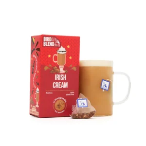 A medium bright red square cardboard box that has Bird & Blend Irish Cream Tea Bag Gift written in bright white writing and some pictures of some creamy white topped drinks and some pieces of dark brown chocolate on it next to a short cylindrical clear glass mug full of some creamy brown liquid and a medium translucent Pyramid shaped Tea Bag, on a white background.