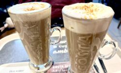 A tall cylindrical clear glass latte glass with a clear glass handle full of some dark brown liquid, some creamy white whipped cream and some pieces of golden brown cookie that has Costa engraved on it next to a tall cylindrical clear glass latte glass with a clear glass handle full of some dark brown liquid, some creamy white whipped cream and some pieces of golden brown cookie that has Costa engraved on it on a large rectangular glossy black tray, on a white background.