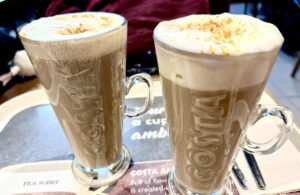 A tall cylindrical clear glass latte glass with a clear glass handle full of some dark brown liquid, some creamy white whipped cream and some pieces of golden brown cookie that has Costa engraved on it next to a tall cylindrical clear glass latte glass with a clear glass handle full of some dark brown liquid, some creamy white whipped cream and some pieces of golden brown cookie that has Costa engraved on it on a large rectangular glossy black tray, on a white background.