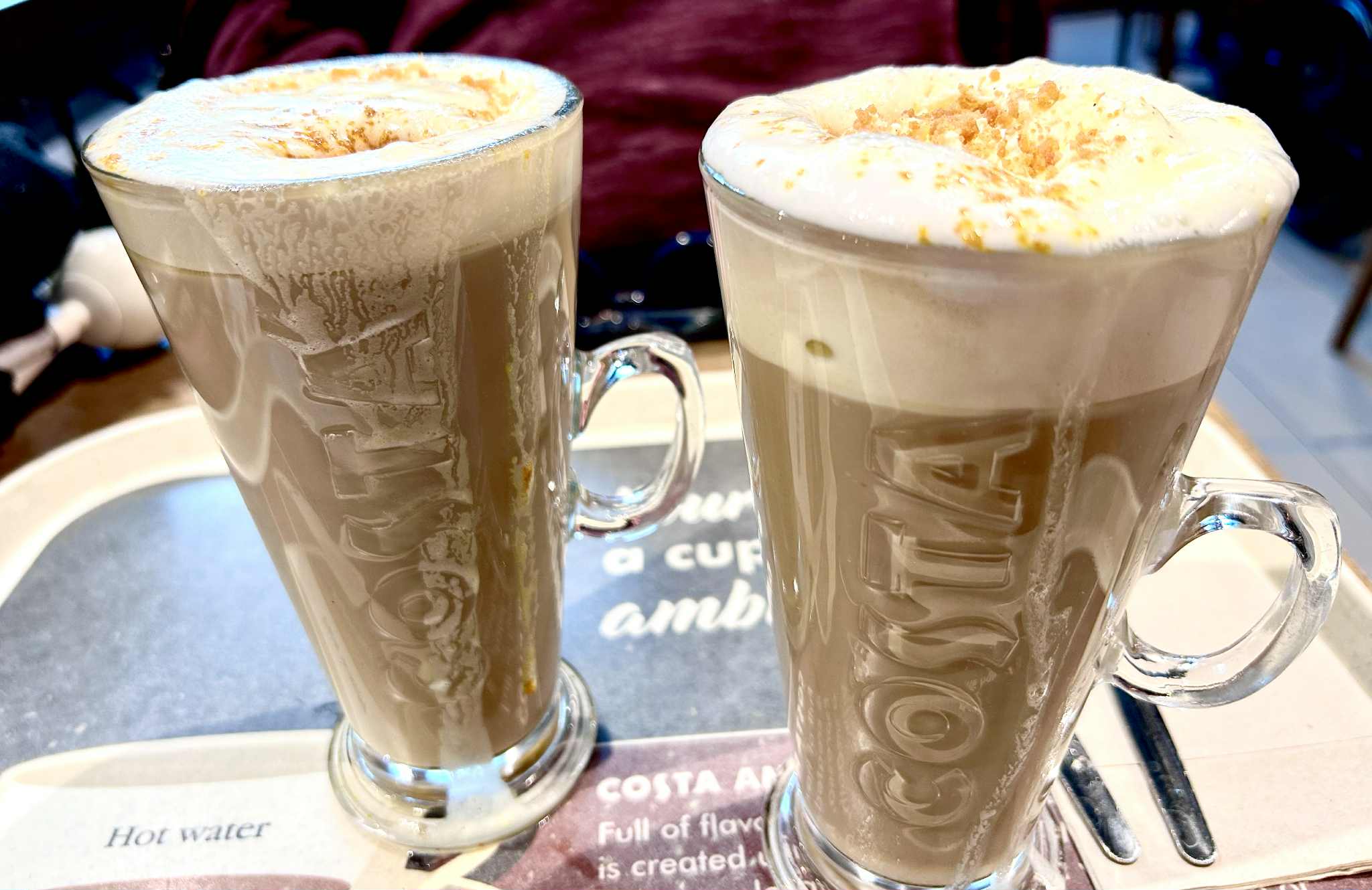 How To Make A Costa Coffee Butter Pecan Cookie Latte At Home A tall cylindrical clear glass latte glass with a clear glass handle full of some dark brown liquid, some creamy white whipped cream and some pieces of golden brown cookie that has Costa engraved on it next to a tall cylindrical clear glass latte glass with a clear glass handle full of some dark brown liquid, some creamy white whipped cream and some pieces of golden brown cookie that has Costa engraved on it on a large rectangular glossy black tray, on a white background.