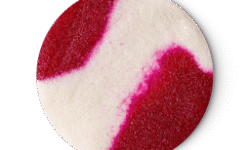A medium circular puddle of some gradient bright magenta red and creamy white body scrub that has some pieces of Castor Sugar running through it, on a light grey background.