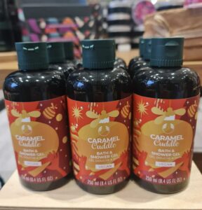 A tall cylindrical dark green plastic bottle with a dark green plastic lid and a gradient bright orange, red and yellow label that has The Body Shop Caramel Cuddle Bath & Shower Gel written in bold black writing and a picture of a swirl of Caramel, a Bee, and some stars on it, on a white background.