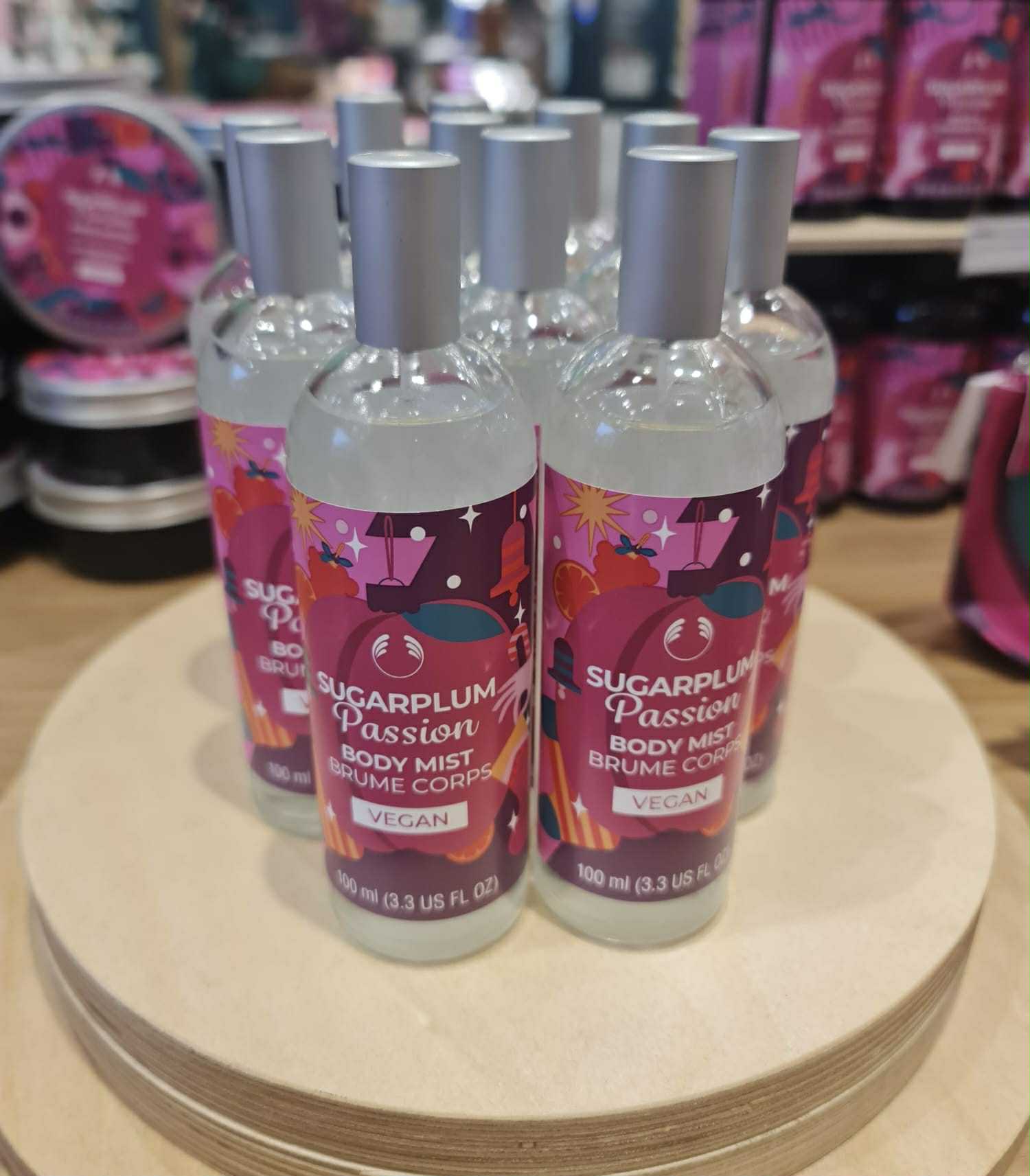 Everyone Deserves A Little Bit Of Passion At Christmas | The Body Shop Christmas 2025 Range A tall cylindrical clear glass bottle full of some clear liquid with a silver metal lid and a gradient bright pink and purple label that has The Body Shop Sugar Pumpkin Body Mist written in bright white writing and a picture of a Christmas Stocking, a Christmas Ornament, some wrapped Christmas Presents, some stars, and some dark red plums on it, on a white background.
