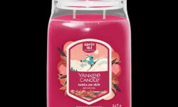 A tall cylindrical glass Jar full of some bright pink Wax with a Label that has Yankee Candle written in bright white writing, Santa On Skis written in bright white writing, and a picture of Santa skiing on some snowy bright white mountains surrounded by some dark green pine trees on it, on a white background.