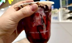 A hand holding a short rounded cylindrical plastic cup full of some creamy white milk, some dark red Cherry Sauce and some dark brown chocolate sauce with a translucent film lid that has Cupp Bubble Tea written in bright white writing on it on a light wooden table.