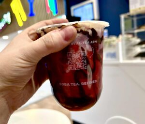 A hand holding a short rounded cylindrical plastic cup full of some creamy white milk, some dark red Cherry Sauce and some dark brown chocolate sauce with a translucent film lid that has Cupp Bubble Tea written in bright white writing on it on a light wooden table.