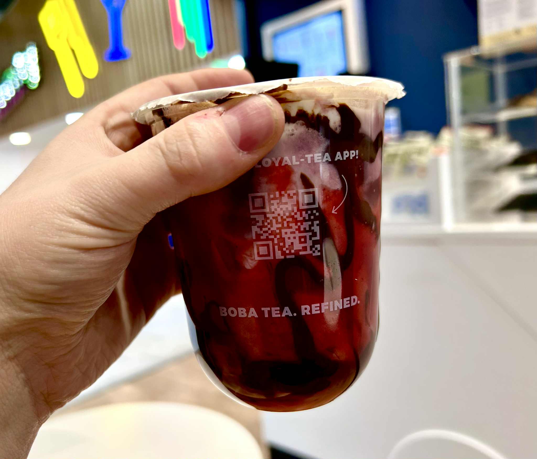 Tea Tastes Terrific | Cupp Bubble Tea X Moomin Range A hand holding a short rounded cylindrical plastic cup full of some creamy white milk, some dark red Cherry Sauce and some dark brown chocolate sauce with a translucent film lid that has Cupp Bubble Tea written in bright white writing on it on a light wooden table.