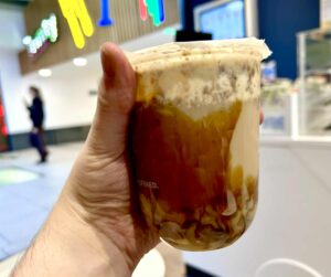 A hand holding a short rounded cylindrical plastic cup full of some creamy white milk and some dark brown Brown Sugar Sauce with a translucent film lid that has Cupp Bubble Tea written in bright white writing on it on a light wooden table?