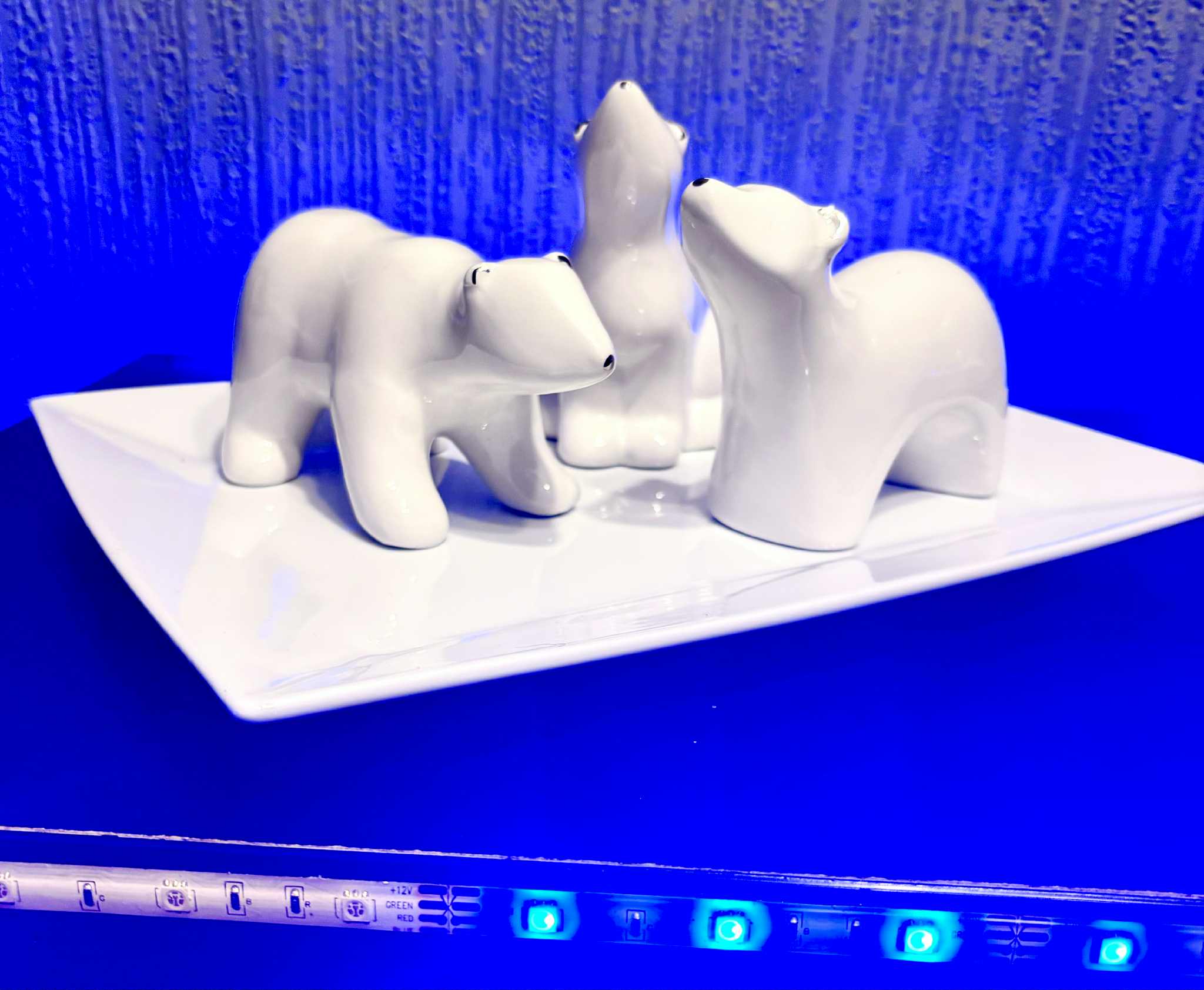 A medium glossy white ceramic Polar Bear Ornament that’s stood on all four Paws, has it’s head slightly downturned as if it’s inspecting something that’s in front of it, some small black eyes, and a small black nose engraved on it next to a medium glossy white ceramic Polar Bear Ornament that has it’s front two paws extended, it’s head tilted upwards as if it’s sniffing the Air, some small black eyes, and a small black nose engraved on it, and a medium glossy white ceramic Polar Bear Ornament that has it’s body slightly curved, it’s head tilted to the left as if it’s looking at something above it, some small black eyes, and a small black nose engraved on it on a large rectangular glossy white plate that’s surrounded by some bright blue fairy lights, on a dark wooden Cabinet.
