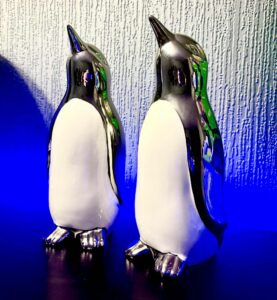 A tall glossy white penguin ornament that has a slightly tilted beak, a metallic silver head, some metallic silver wings, and some metallic silbver feet next to a tall glossy white penguin ornament that has a slightly tilted beak, a metallic silver head, some metallic silver wings, and some metallic silbver feet surrounded by some bright blue fairy lights, on a dark wooden cabinet.