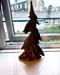 A large dark brown resin Christmas tree ornament that has some textured layers all over it that look like branches on a glossy white windowsill in front of a large rectangular window looking out on to some dark stone buildings.