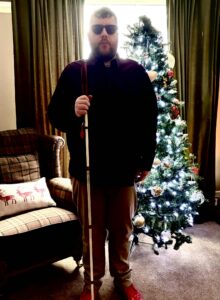 A tall bearded man wearing a pair of dark denim blue glasses with some pale red lenses, a dark red long sleeve Oxford Shirt, a burgundy red crew neck t-shirt, a pair of light mushroom grey chino trousers and a pair of shiny bright red shoes holding a long bright white cane with a bright red leather handle stood in front of a large dark green Christmas tree.