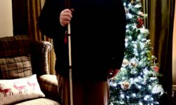 A tall bearded man wearing a pair of dark denim blue glasses with some pale red lenses, a dark red long sleeve Oxford Shirt, a burgundy red crew neck t-shirt, a pair of light mushroom grey chino trousers and a pair of shiny bright red shoes holding a long bright white cane with a bright red leather handle stood in front of a large dark green Christmas tree.