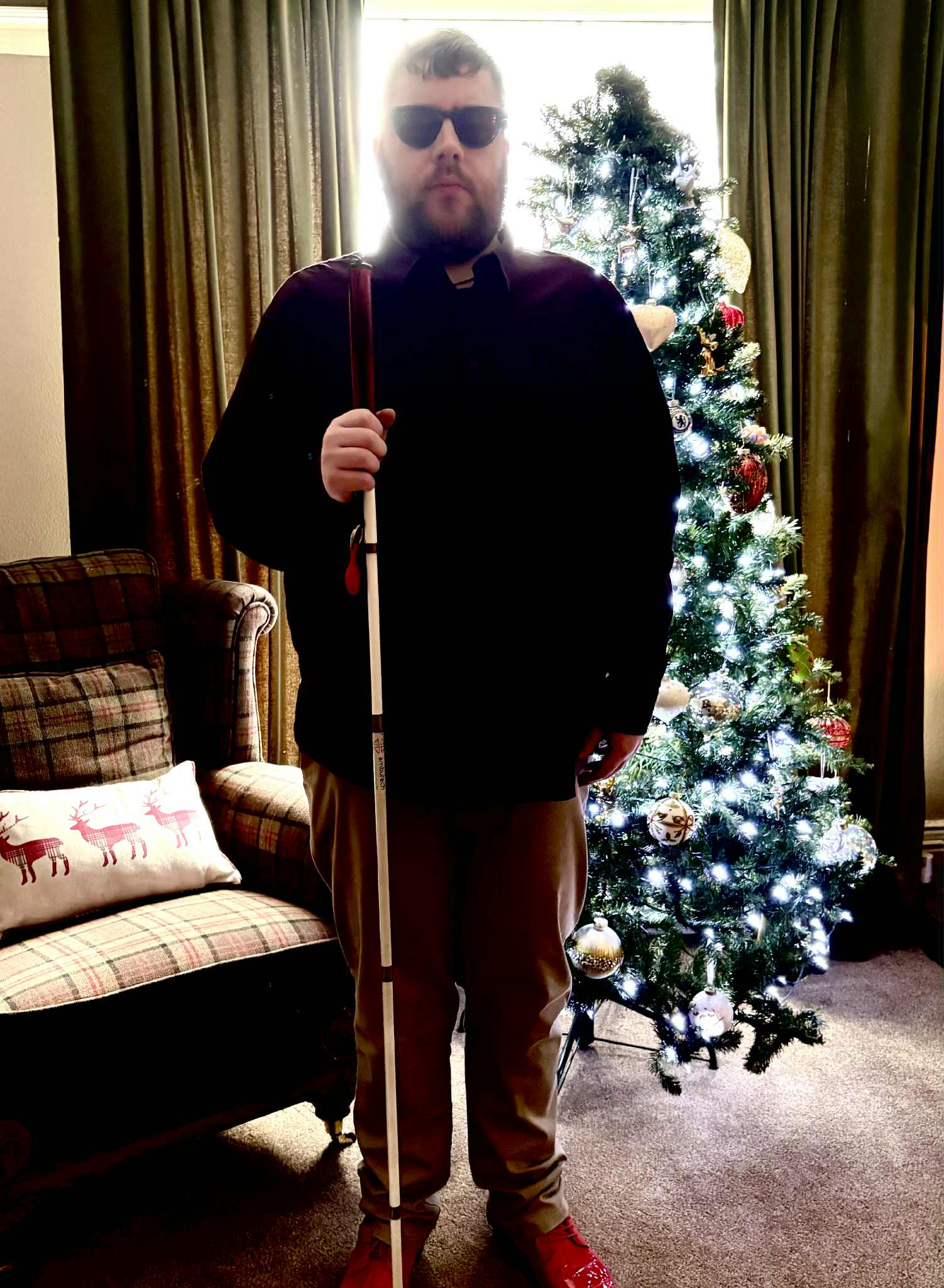 Outfit Of The Day | Christmas Cheer Is All Around Us This Year A tall bearded man wearing a pair of dark denim blue glasses with some pale red lenses, a dark red long sleeve Oxford Shirt, a burgundy red crew neck t-shirt, a pair of light mushroom grey chino trousers and a pair of shiny bright red shoes holding a long bright white cane with a bright red leather handle stood in front of a large dark green Christmas tree.