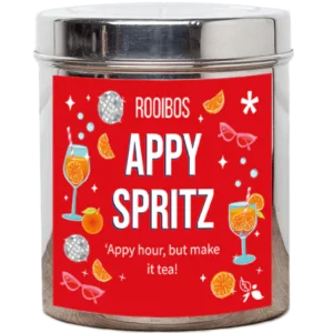 A tall cylindrical bright silver metal tin with a bright silver lid and a bright red label that has Apy Spritz loose Leaf Tea written in bright white writing and some pictures of some cocktail glasses garnished with some slices of orange, some sparkling disco balls, some slices of Orange, some stars, some bright red sunglasses, and some bright white dots on it, on a white background.