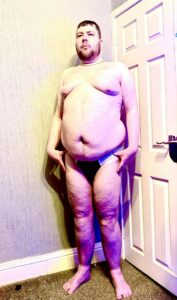 A tall bearded man with his hands on his hips wearing a pair of black briefs that has some white trim all over the seems and a white waistband that has M:R Underwear written in bold black writing on it stood against a white wooden door.