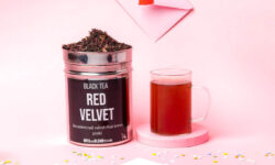 A tall cylindrical bright silver metal tin full of some tea leaves with a bright silver lid and a black label that has Red Velvet loose Leaf Tea written in bright white writing on it next to a tall cylindrical clear glass Mug full of some bright red liquid on top of a bright pink round shaped coaster and a large rectangular white envelope that has a bright red love heart seal attached to it surrounded by some multicoloured confetti, on a bright pink background.