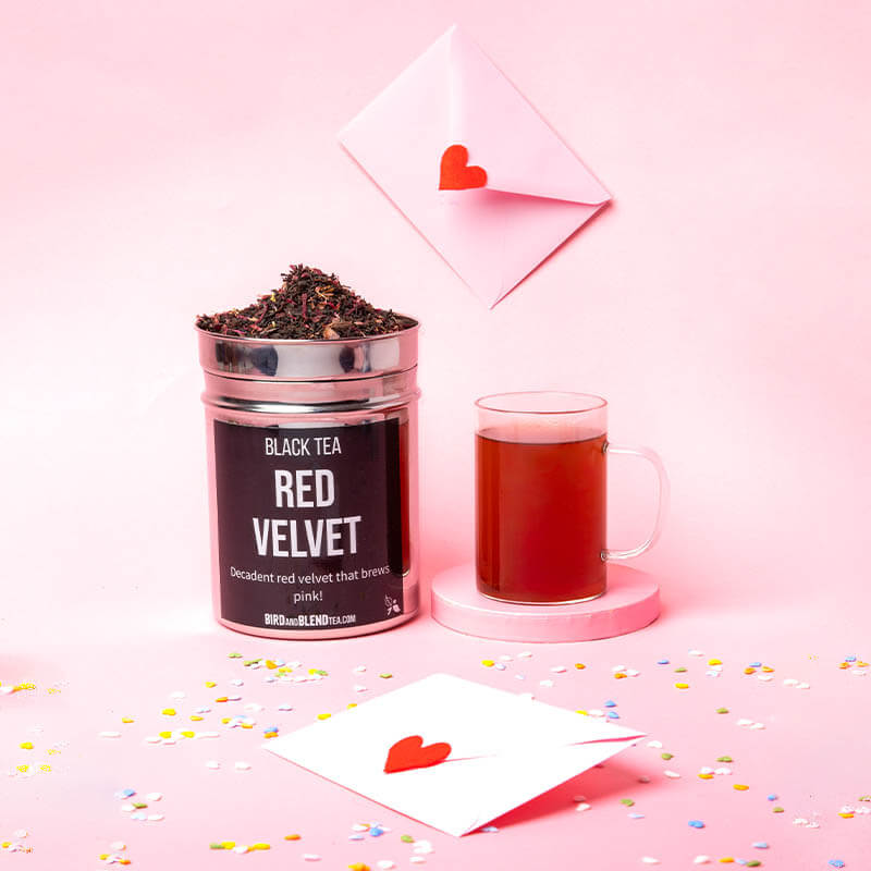 Tea Tastes Terrific | Bird & Blend Tea Company Valentine's Day 2026 Collection A tall cylindrical bright silver metal tin full of some tea leaves with a bright silver lid and a black label that has Red Velvet loose Leaf Tea written in bright white writing on it next to a tall cylindrical clear glass Mug full of some bright red liquid on top of a bright pink round shaped coaster and a large rectangular white envelope that has a bright red love heart seal attached to it surrounded by some multicoloured confetti, on a bright pink background.