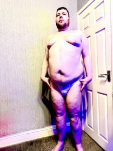 A tall bearded man looking slightly upwards with his hands on his hips wearing a bright white tanga brief with a black waistband that has Kiniki written in bright white writing on it stood against a white wooden door.