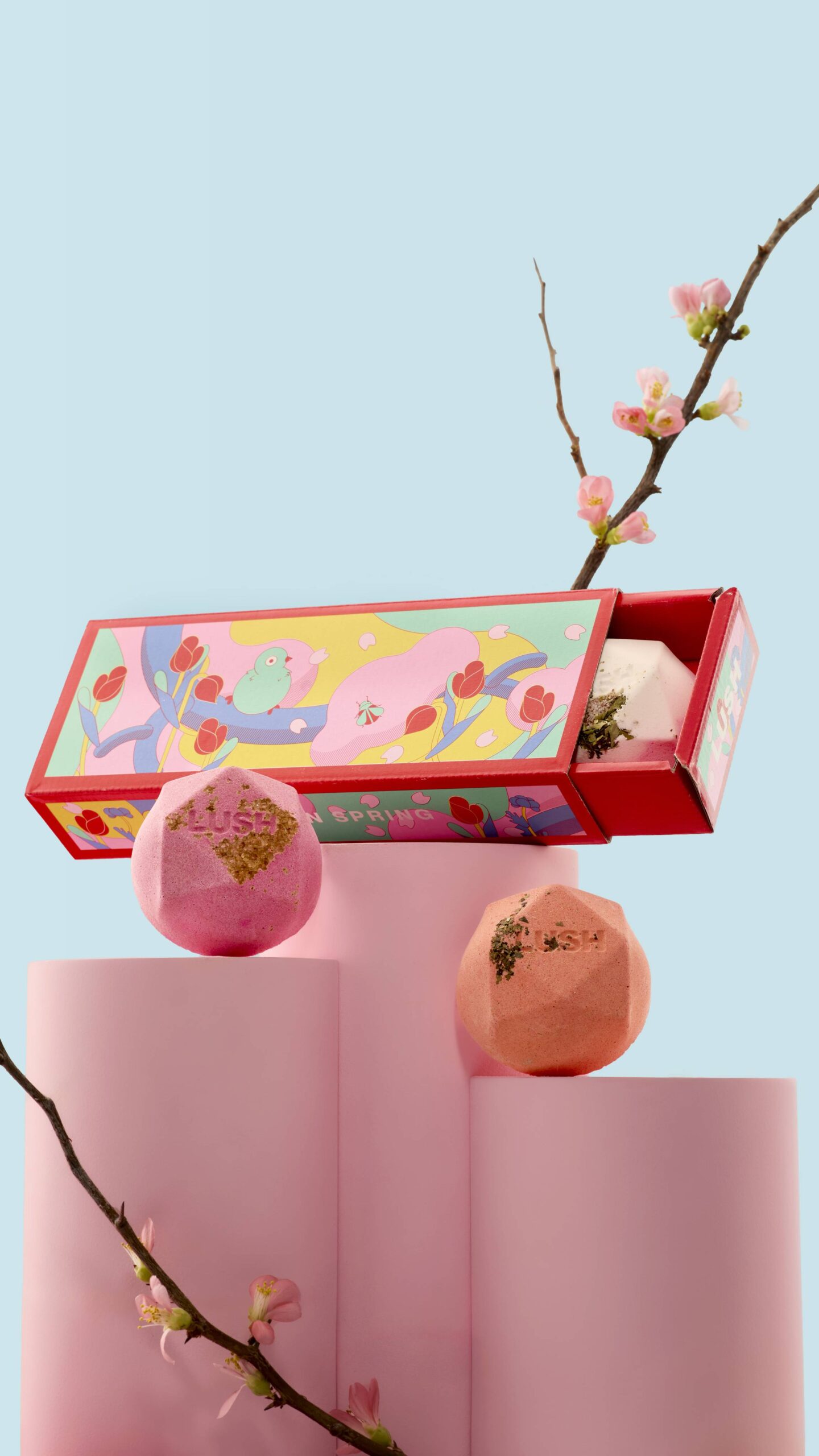 There Will Be No White Flag Above My Door. I'm In Love And Always Will Be | Lush White Day 2026 Range A small bright pink flat hexagonal shaped bath bomb next to a small pale pink flat hexagonal shaped bath bomb, and a A small bright white flat hexagonal shaped bath bomb that has some pieces of dried bright pink and red petals embedded in it inside a long and thin bright red cardboard tray with a bright turquoise sleeve that has a bright yellow boarder and some pictures of a light blue bird, some bright red tulips, some bright green leaves, and some stylised bright pink shapes on it, on a white background.