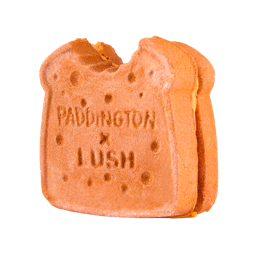 A large dark orange square sandwich shaped bubble bar that has some creamy white Cocoa Butter sandwiched in between it, a small bite taken out of the corner of it, and Lush x Paddington engraved on it, on a white background.