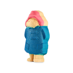 Take It Away, Paddington! | Lush X Paddington™ Range | Gifted A medium dark tan brown bear shaped bath bomb with a bright red hat and a dark blue raincoat that has a neutral expression engraved on it and some bright gold lustre all over it, on a white background.