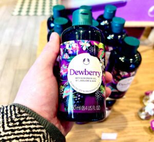A hand holding a tall cylindrical dark green plastic Bottle with a dark green plastic lid and a dark purple label that has The Body Shop Dewberry Bath & Shower Gel written in dark purple writing and some pictures of some small dark purple blackberries, some bright white flowers, and some dark green leaves on it, on a white background.