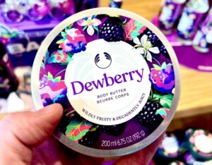 A hand holding a short cylindrical dark green plastic tub with a dark green plastic lid and a bright white label that has The Body Shop Dewberry Body Butter written in dark purple writing and some pictures of some small dark purple blackberries, some bright white flowers, and some dark green leaves on it, on a white background.