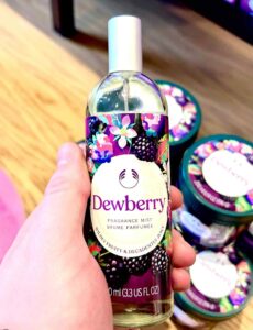 A hand holding a tall cylindrical clear glass bottle full of some clear liquid with a silver metal lid and a dark purple label that has The Body Shop Dewberry Fragrance Mist written in dark purple writing and some pictures of some dark purple blackberries, some bright white flowers, and some dark green leaves on it, on a white background.