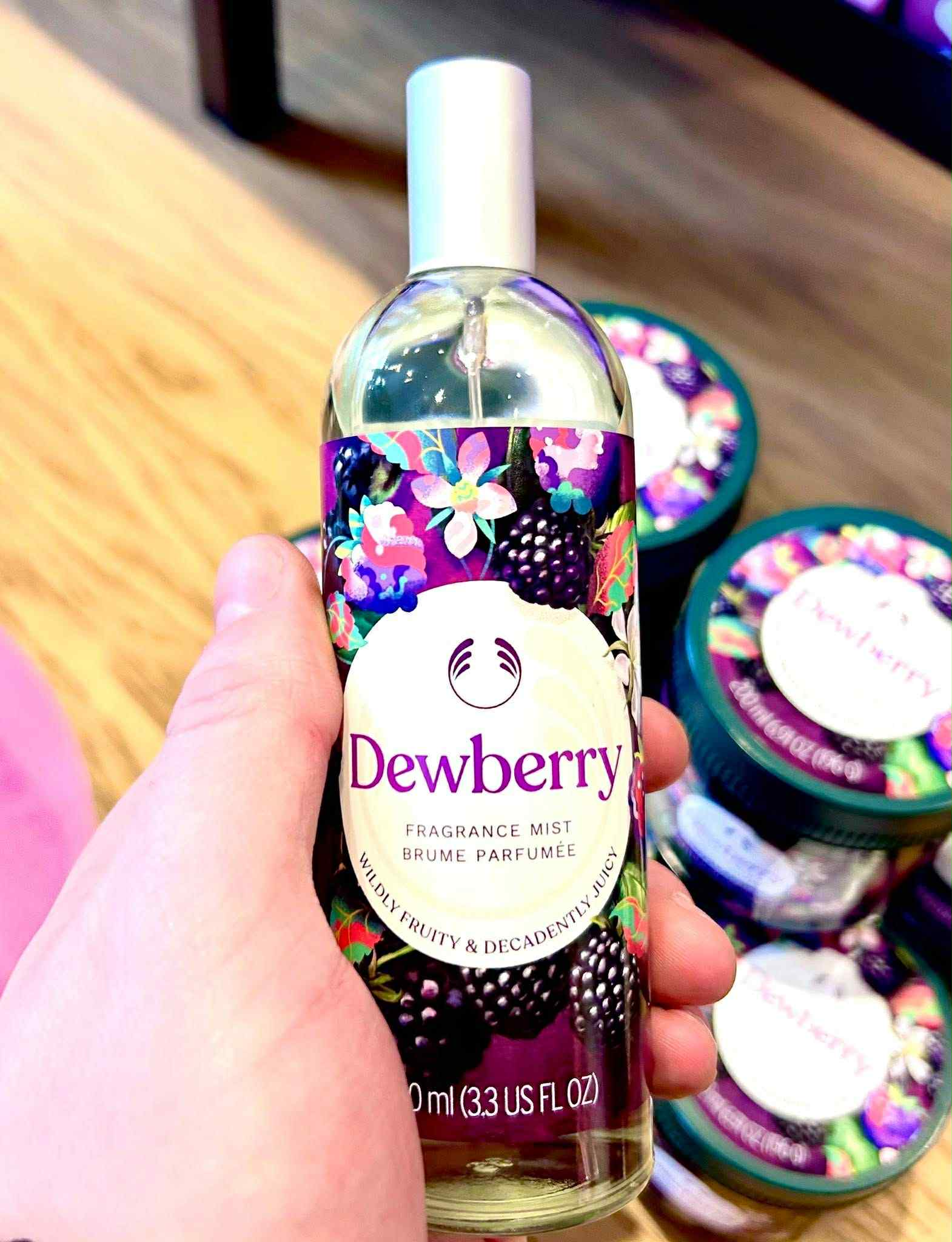 A hand holding a tall cylindrical clear glass bottle full of some clear liquid with a silver metal lid and a dark purple label that has The Body Shop Dewberry Fragrance Mist written in dark purple writing and some pictures of some dark purple blackberries, some bright white flowers, and some dark green leaves on it, on a white background.