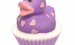 A large circular bright white cupcake shaped block of soap that has a pale Lilac paper cupcake case around it and a bright purple Rubber Duck with a bright orange beak, some large eyes, and some bright pink heart shapes all over it sat on top of it, on a white background.