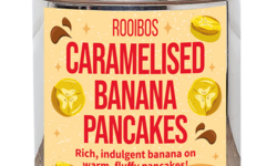 A tall cylindrical bright silver metal tin with a bright silver lid and a pale yellow label that has Caramelised Banana Pancakes loose Leaf Tea written in bright red writing and some pictures of some slices of bright yellow bananas and some dark brown puddles of Caramel Sauce on it, on a white background.