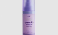 A short cylindrical bright Purple Bottle with a bright purple pump lid that’s covered by a matte semi-translucent purple plastic cap lid that has Primark PS… Glass Skin Face Serum written in dark purple writing on it, on a white background.