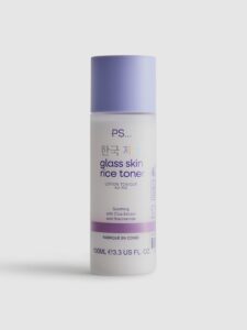 A tall cylindrical frosted white plastic bottle with a pale lavender plastic lid that has Primark PS… Glass Skin Rice Face Toner written in bright Purple writing on it, on a white background.
