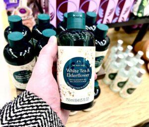 A hand holding a tall cylindrical dark green plastic Bottle with a bright teal plastic lid and a bright white label that has The Body Shop White Tea & Elderflower Bath & Shower Gel written in bright white writing inside a dark teal circle and some pictures of some small bright white blossoms with some pale yellow Pistils and some bright green stems on it, on a white background.