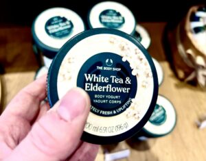 A hand holding a short rounded rectangular dark green plastic tub with a bright teal plastic lid and a bright white label that has The Body Shop White Tea & Elderflower Body Yoghurt written in bright white writing inside a dark teal circle and some pictures of some small bright white blossoms with some pale yellow {Pistils and some bright green stems on it, on a white background.