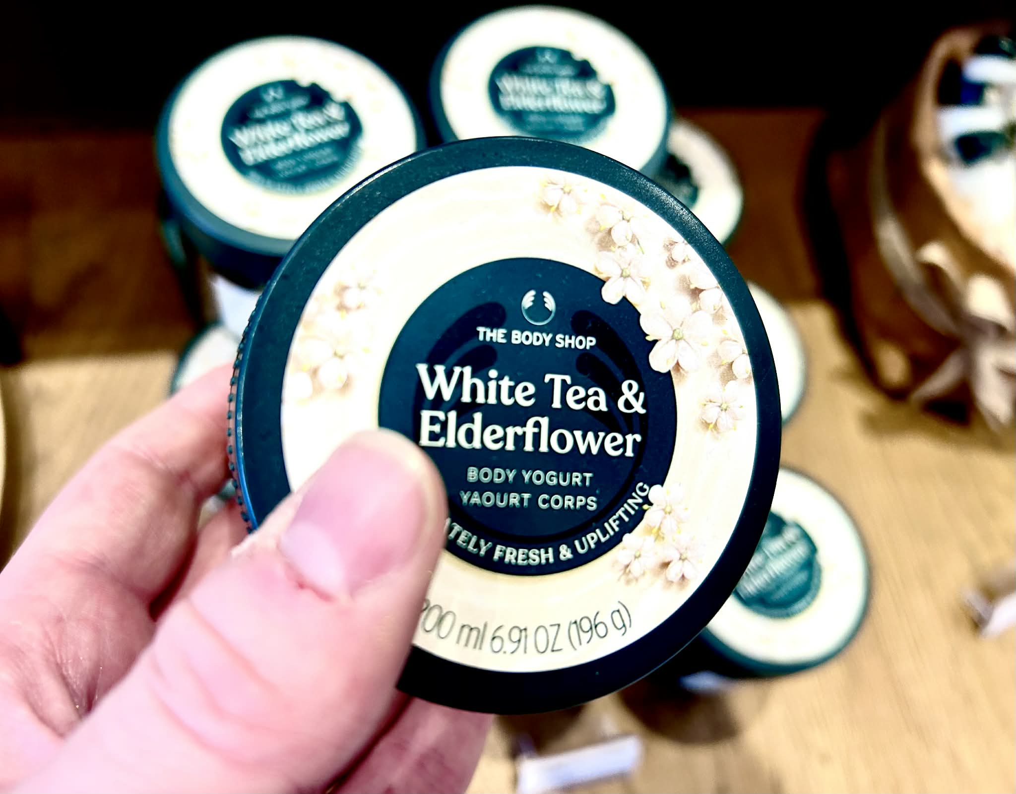 A hand holding a short rounded rectangular dark green plastic tub with a bright teal plastic lid and a bright white label that has The Body Shop White Tea & Elderflower Body Yoghurt written in bright white writing inside a dark teal circle and some pictures of some small bright white blossoms with some pale yellow {Pistils and some bright green stems on it, on a white background.