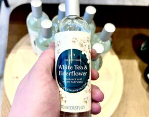 A hand holding a tall cylindrical clear glass bottle full of some clear liquid with a white metallic lid and a bright teal label that has The Body Shop White Tea & Elderflower Fragrance Mist written in bright white writing inside a dark teal circle and some pictures of some small bright white blossoms with some pale yellow Pistels and some bright green stems on it, on a white background.