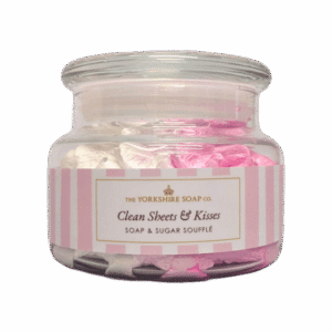 A short cylindrical clear glass jar full of some gradient bright pink and creamy white liquid that has some pieces of Sugar running through it with a clear flared glass lid and a gradient bright pink and white label that has The Yorkshire Soap Company Clean Sheets & Kisses Soap & Sugar Soufflé written in bold black writing and a picture of a black crown on it, on a white background.