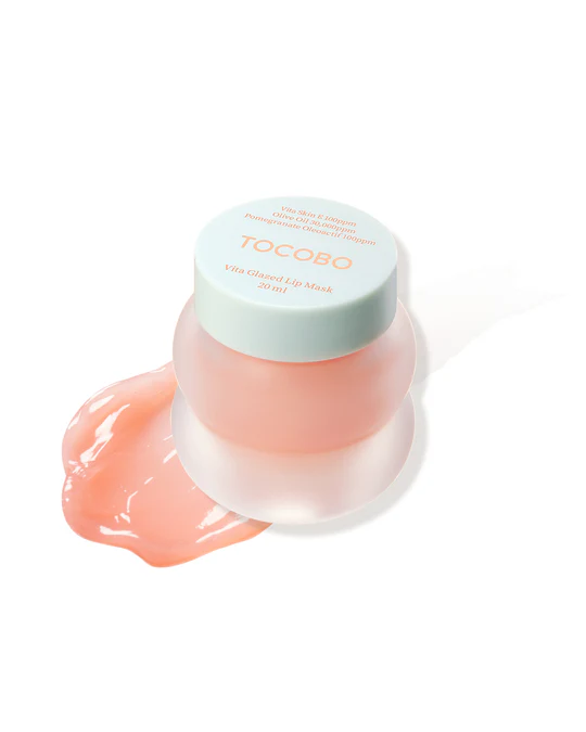 Getting A Bit Of Glaze On Your Lips Is Actually Helping Them | Tocobo Vita Glazed Lip Mask A short cylindrical frosted Peach pink glass jar with a Pale mint green plastic lid that has Tocobo Vita Glazed Lip Mask written in pale orange writing on it next to a large swatch of some translucent pale pink lip mask, on a white background.