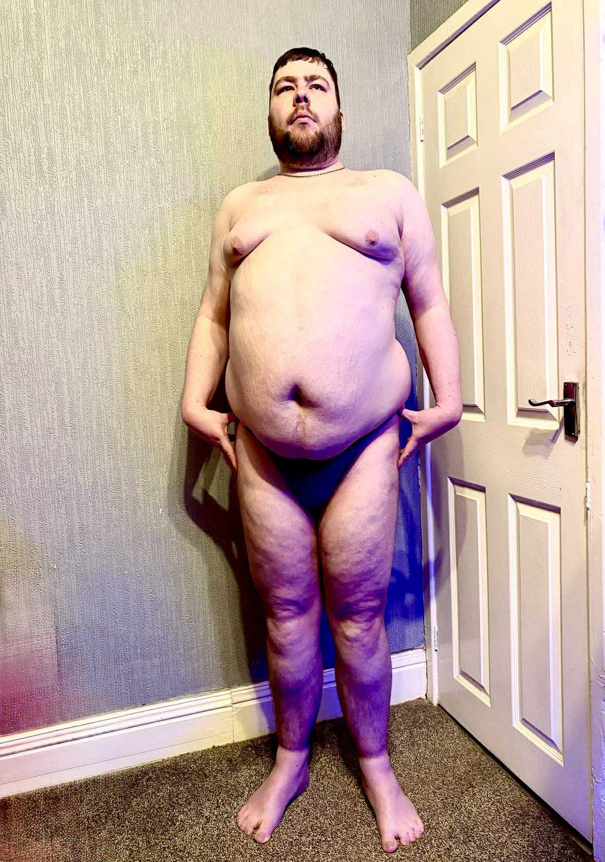 A tall bearded man with his hands on his hips wearing a dark turquoise tanga with a dark turquoise waistband that has Kiniki written in bold white writing on it stood in front of a white wooden door.